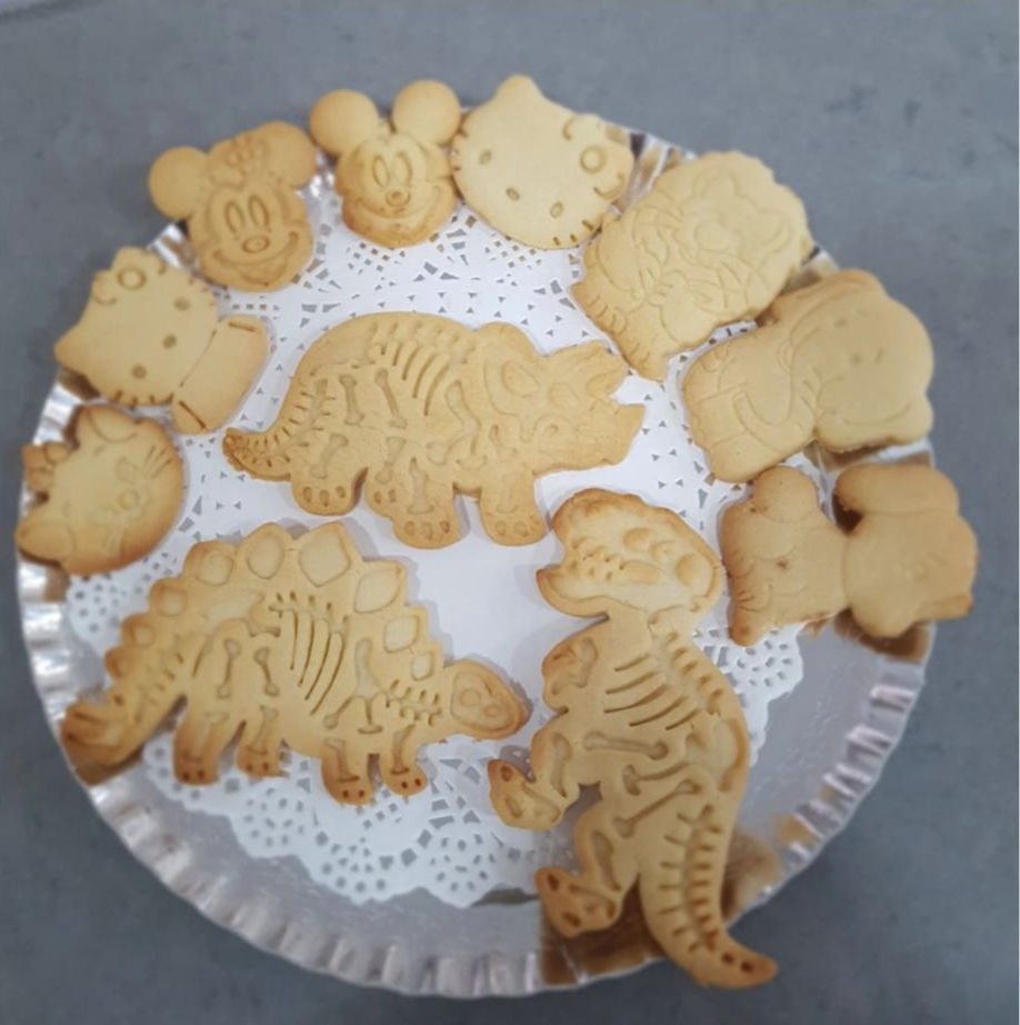 A round tray of playful cookies shaped like dinosaurs and popular cartoon characters including Hello Kitty, Mickey Mouse, and Snoopy, placed on a paper doily.